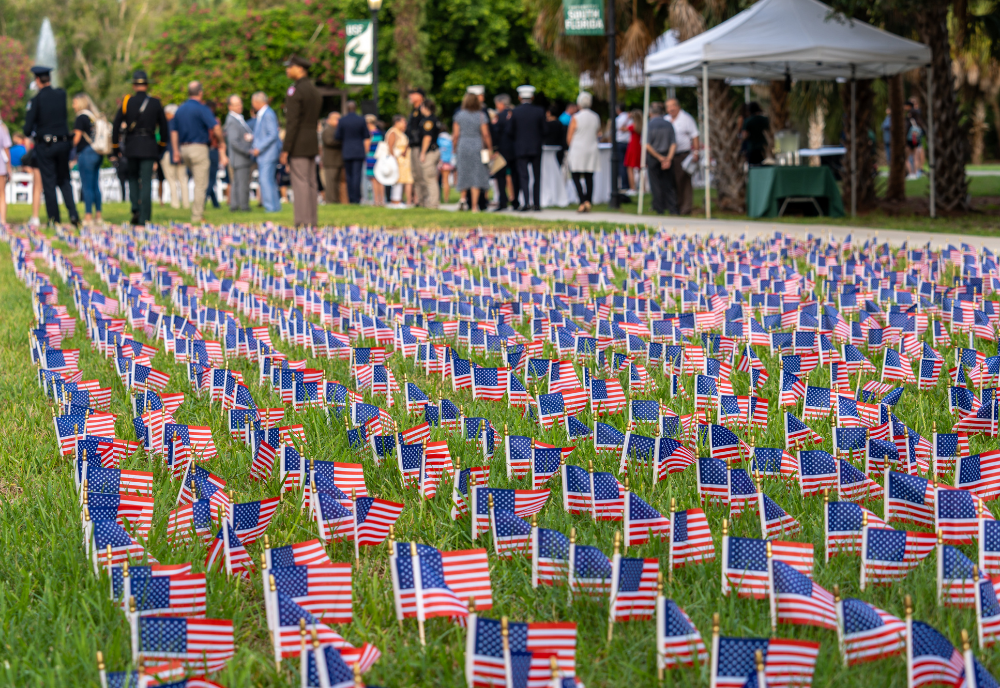 Hundreds of small American flags displayed in rows on a grassy lawn, with people gathered in the background near a white tent for a remembrance ceremony.