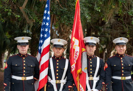 Four U.S. Marines in dress uniforms stand at attention outdoors, holding the American flag and the Marine Corps flag.