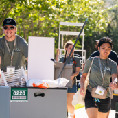 A group of USF volunteers wearing green shirts help students move into campus housing by pushing carts filled with belongings along a sunny outdoor pathway.
